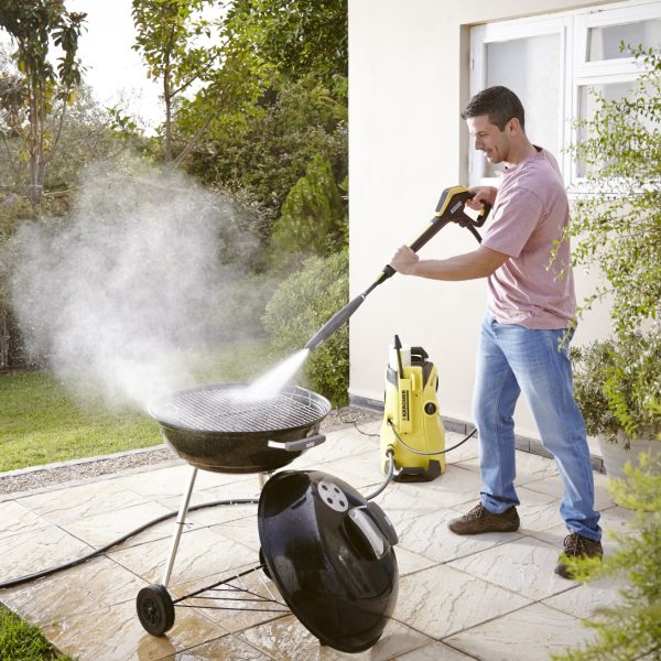 Man using a K4 Power Control Home Pressure Washer to clean a dirty barbecue grill in a backyard.