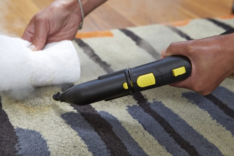 A person uses a Kärcher SC 2 Easyfix Steam Cleaner on a striped carpet, with a focus on the device and the cleaning action.